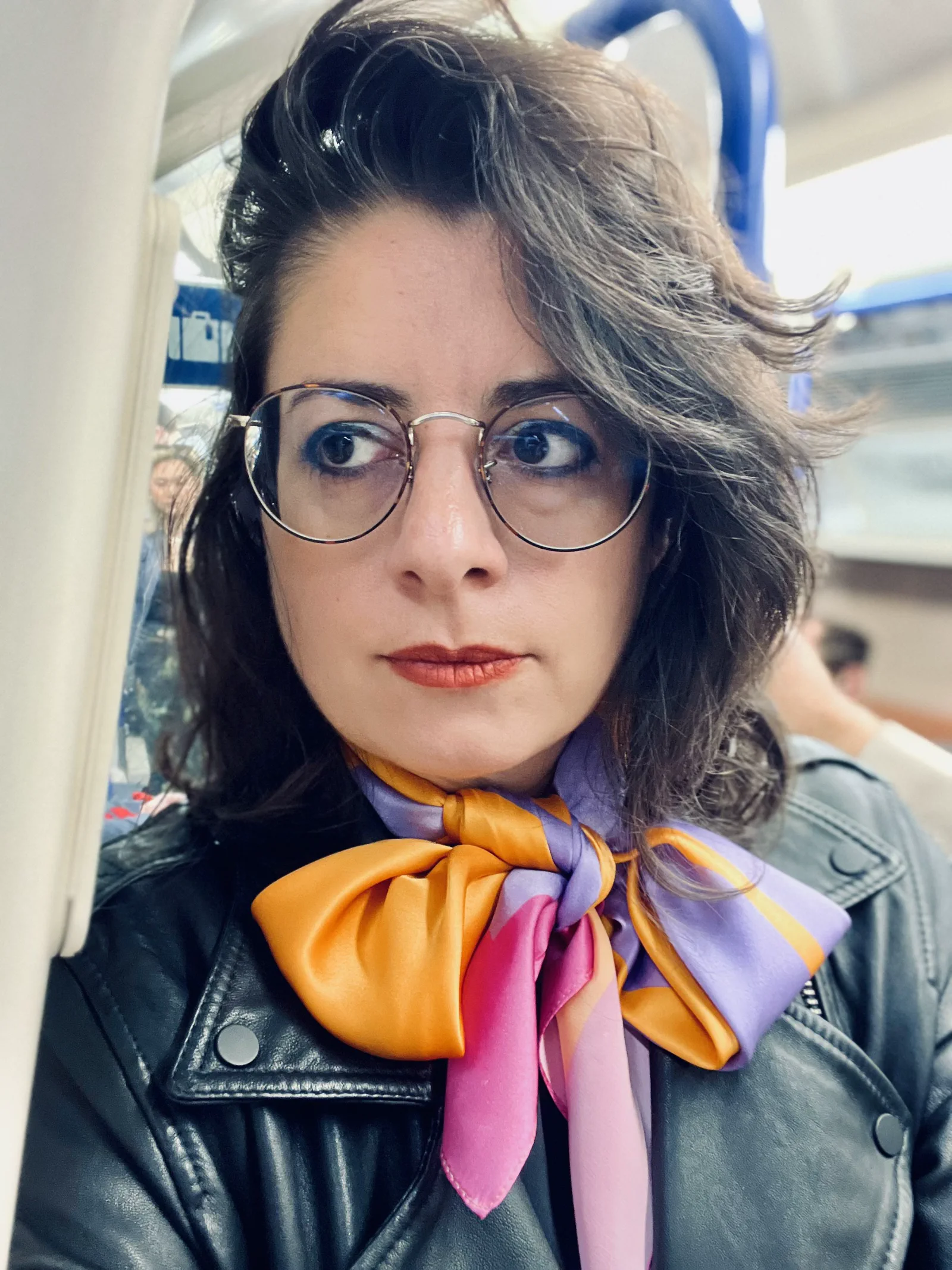 Contemporary patternist Kika Pierides wearing round glasses, a black leather jacket, and a colourful orange, pink and purple silk scarf of her own textile design