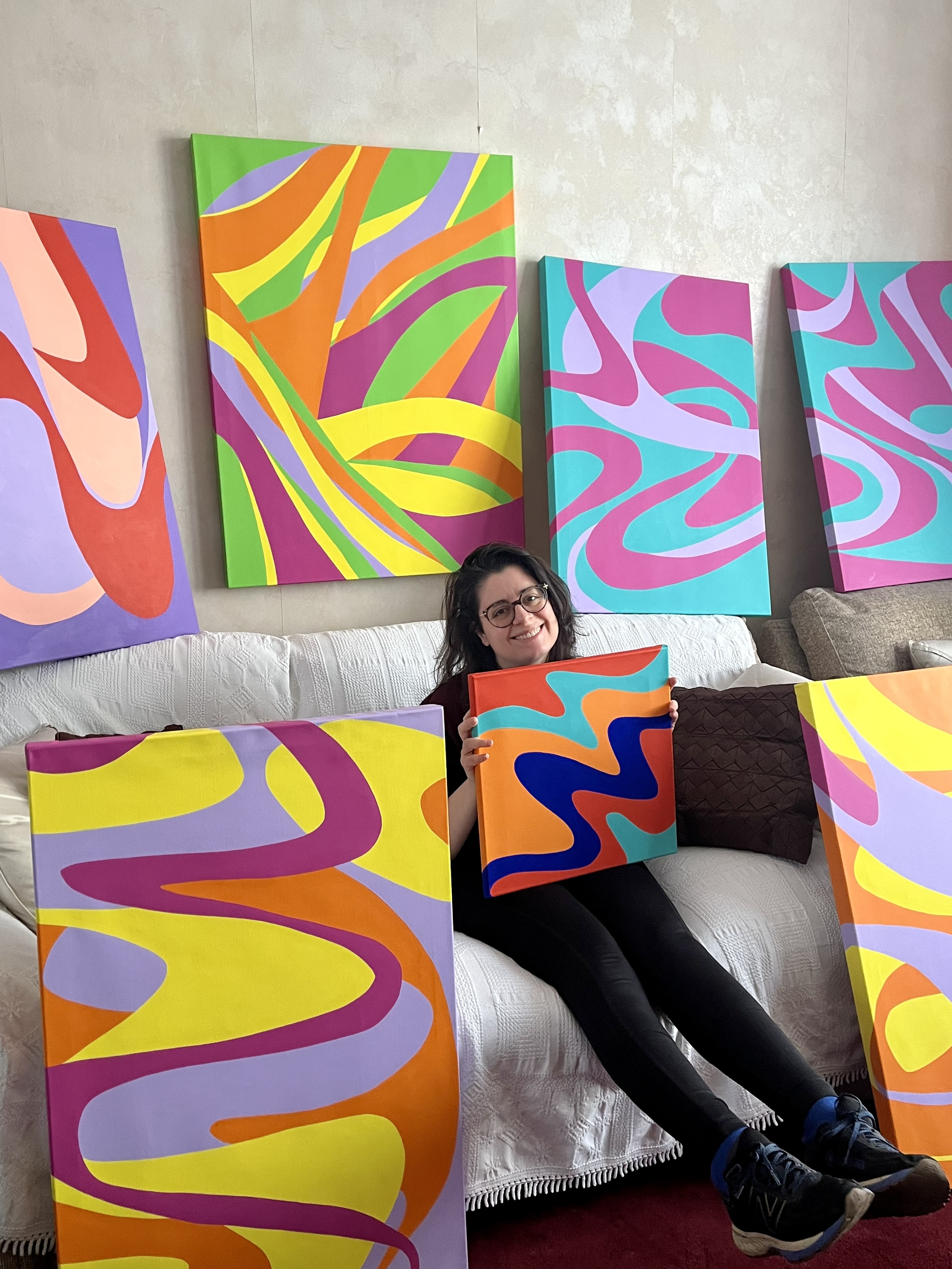 Contemporary patternist and textile artist Kika Pierides sitting on a couch, holding a colourful abstract painting, with more paintings leaning against the couch and hanging on the wall behind her