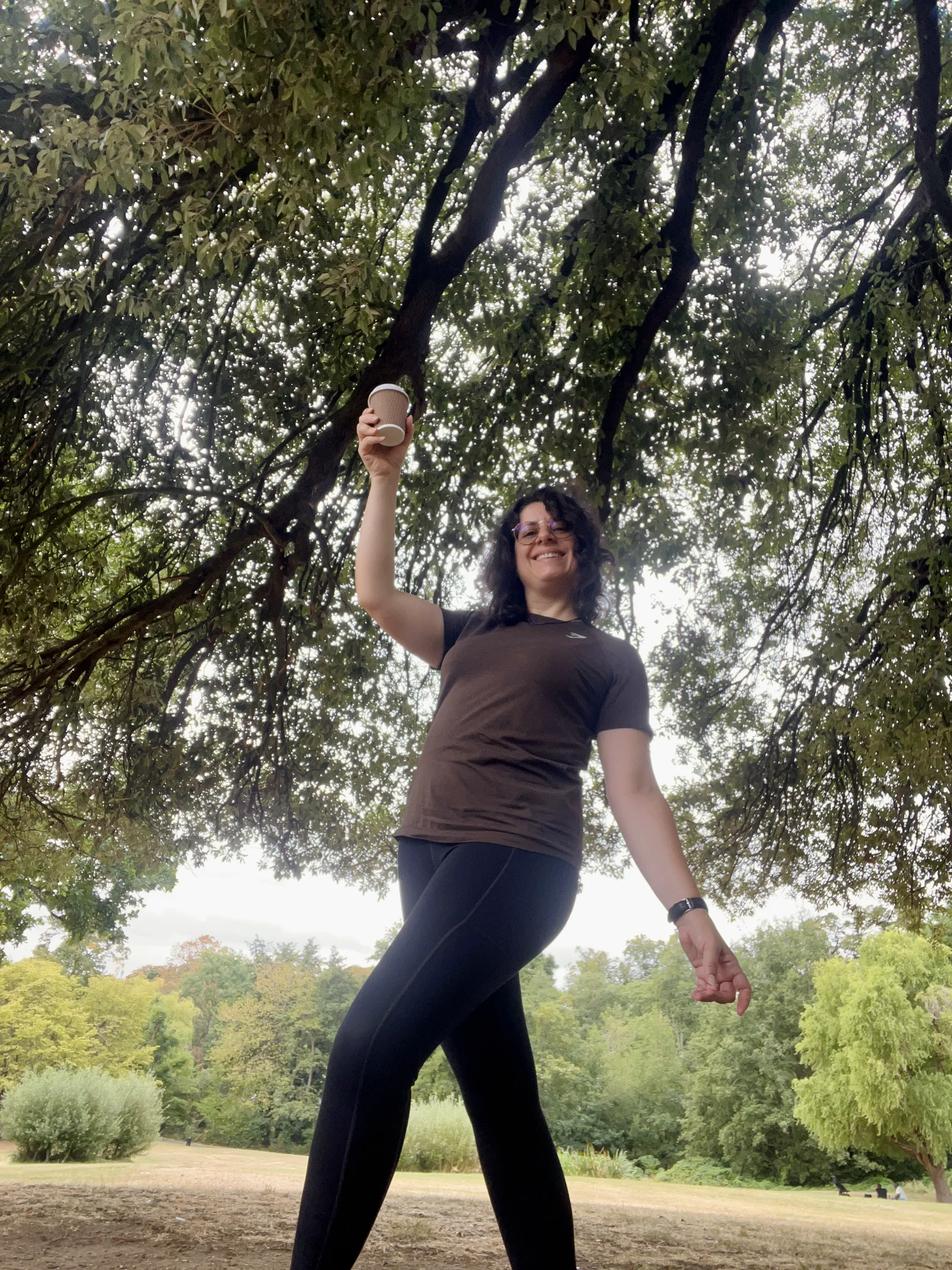Kika Pierides raising a coffee cup beneath the trees in a park.