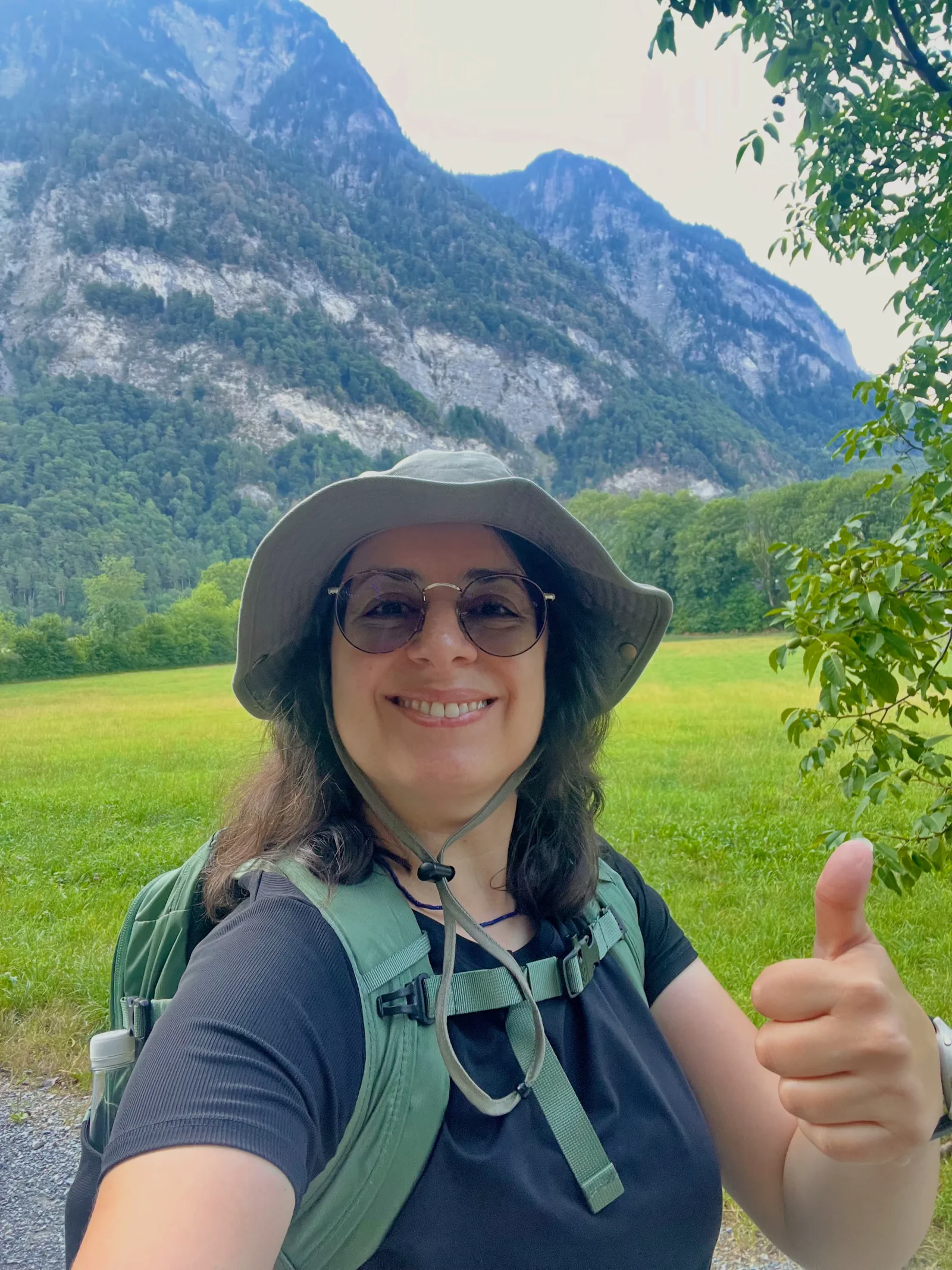 Kika Pierides hiking in Liechtenstein in front of a mountain, wearing a hat and giving a thumbs-up.